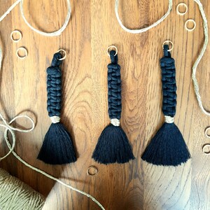 Op de afbeelding: Drie zwarte macramé sleutelhangers met kwastjes die aan een houten oppervlak hangen. De sleutelhangers zijn gemaakt van zwart touw en hebben een lichtbruin kwastje aan de onderkant.