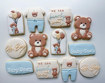 May include: A collection of baby shower-themed sugar cookies. The cookies are decorated with brown, white, and blue icing, featuring teddy bears, balloons, and text that reads "Oh Boy", "Baby Goetz", and "We can bearly wait".