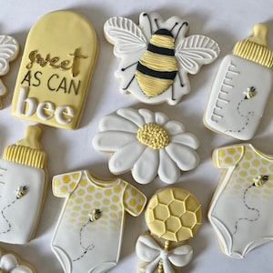 K&ouml;nnte beinhalten: Ein Set mit 10 dekorierten Zuckerkeksen mit Bienen-Motiv. Die Kekse sind mit gelbem, wei&szlig;em und schwarzem Zuckerguss verziert. Die Kekse beinhalten eine Biene, eine G&auml;nsebl&uuml;mchen, eine Babyflasche, einen Strampler und eine Rassel. Die Kekse sind mit dem Text "sweet as can bee" verziert.