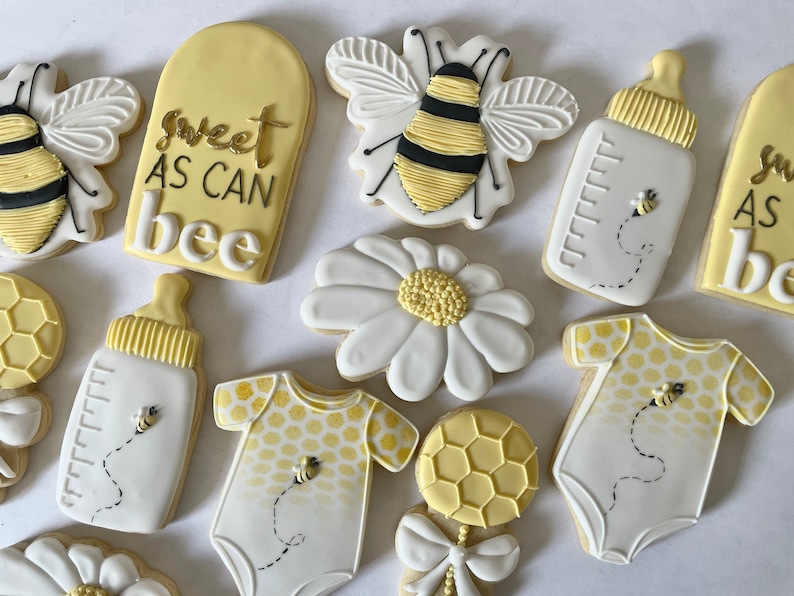 K&ouml;nnte beinhalten: Ein Set mit 10 dekorierten Zuckerkeksen mit Bienen-Motiv. Die Kekse sind mit gelbem, wei&szlig;em und schwarzem Zuckerguss verziert. Die Kekse beinhalten eine Biene, eine G&auml;nsebl&uuml;mchen, eine Babyflasche, einen Strampler und eine Rassel. Die Kekse sind mit dem Text "sweet as can bee" verziert.