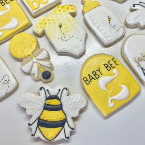 K&ouml;nnte beinhalten: Ein Set aus neun gelben und wei&szlig;en, verzierten Zuckerkeksen mit Bienen- und Babymotiven. Die Kekse zeigen eine Babyflasche, eine Rassel, einen Strampler, eine Biene und Schilder mit der Aufschrift "Baby Bee" und "Sweet as can bee".