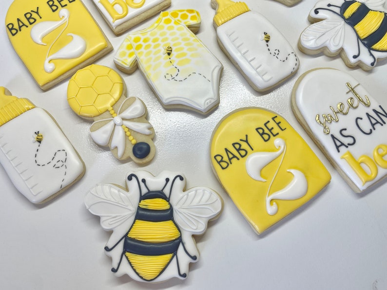 K&ouml;nnte beinhalten: Ein Set aus neun gelben und wei&szlig;en, verzierten Zuckerkeksen mit Bienen- und Babymotiven. Die Kekse zeigen eine Babyflasche, eine Rassel, einen Strampler, eine Biene und Schilder mit der Aufschrift "Baby Bee" und "Sweet as can bee".