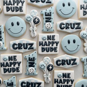 May include: A collection of light blue and white decorated sugar cookies featuring the text ONE HAPPY DUDE, the name CRUZ, the number 1, smiley faces, and balloon clusters with lightning bolt designs, all rendered in black icing on a light background.