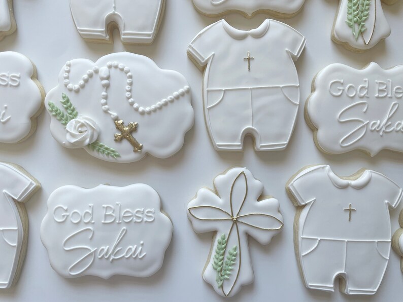 Pode incluir: Biscoitos de a&ccedil;&uacute;car branco decorados com glac&ecirc; real. Os biscoitos apresentam uma cruz, um ros&aacute;rio, um macac&atilde;o de beb&ecirc; e o texto "God Bless Sakai".