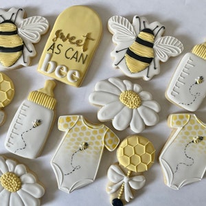 K&ouml;nnte beinhalten: Ein Set mit 10 dekorierten Zuckerkeksen mit Bienen-Motiv. Die Kekse sind mit gelbem, wei&szlig;em und schwarzem Zuckerguss verziert. Die Kekse beinhalten eine Biene, eine Babyflasche, eine Rassel, eine G&auml;nsebl&uuml;mchen und einen Strampler. Die Kekse sind mit dem Text "Sweet as can bee" verziert.