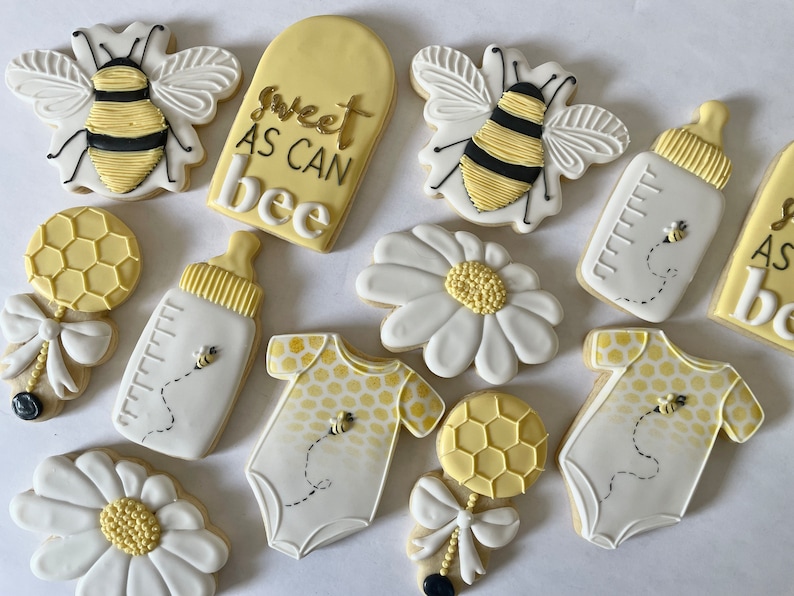 K&ouml;nnte beinhalten: Ein Set mit 10 dekorierten Zuckerkeksen mit Bienen-Motiv. Die Kekse sind mit gelbem, wei&szlig;em und schwarzem Zuckerguss verziert. Die Kekse beinhalten eine Biene, eine Babyflasche, eine Rassel, eine G&auml;nsebl&uuml;mchen und einen Strampler. Die Kekse sind mit dem Text "Sweet as can bee" verziert.