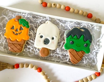 May include: Three decorated sugar cookies in a white box with white paper shred. One cookie is an orange pumpkin ice cream cone, one is a white ghost popsicle, and one is a green Frankenstein monster ice cream cone with a brown waffle pattern.