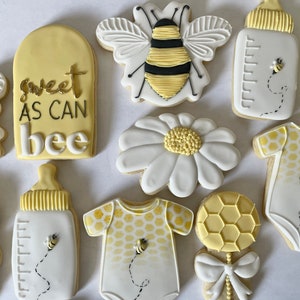 K&ouml;nnte beinhalten: Ein Set aus neun dekorierten Zuckerkeksen mit Bienen-Motiv. Die Kekse sind mit gelbem, wei&szlig;em und schwarzem Zuckerguss verziert. Die Kekse beinhalten eine Biene, eine G&auml;nsebl&uuml;mchen, eine Babyflasche, einen Strampler und eine Rassel. Ein Keks tr&auml;gt den Text "sweet as can bee".