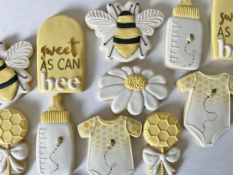 K&ouml;nnte beinhalten: Ein Set aus neun dekorierten Zuckerkeksen mit Bienen-Motiv. Die Kekse sind mit gelbem, wei&szlig;em und schwarzem Zuckerguss verziert. Die Kekse beinhalten eine Biene, eine G&auml;nsebl&uuml;mchen, eine Babyflasche, einen Strampler und eine Rassel. Ein Keks tr&auml;gt den Text "sweet as can bee".