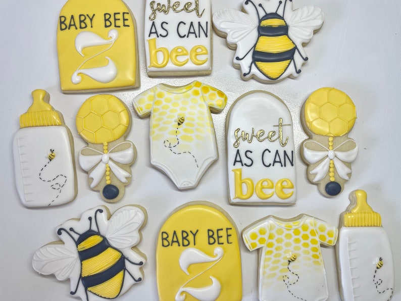 K&ouml;nnte beinhalten: Ein Set aus neun gelben und wei&szlig;en, verzierten Zuckerkeksen mit Bienen- und Babymotiven. Die Kekse zeigen Bilder von Bienen, Babyflaschen, Stramplern und den Text "Baby Bee" und "Sweet as can bee".