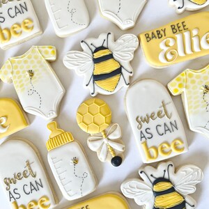 K&ouml;nnte beinhalten: Ein Set mit 10 gelben und wei&szlig;en, verzierten Zuckerkeksen mit Bienen- und Babymotiven. Die Kekse zeigen den Text "Sweet as can bee", "Baby Bee Ellie" und "Bee Ellie".