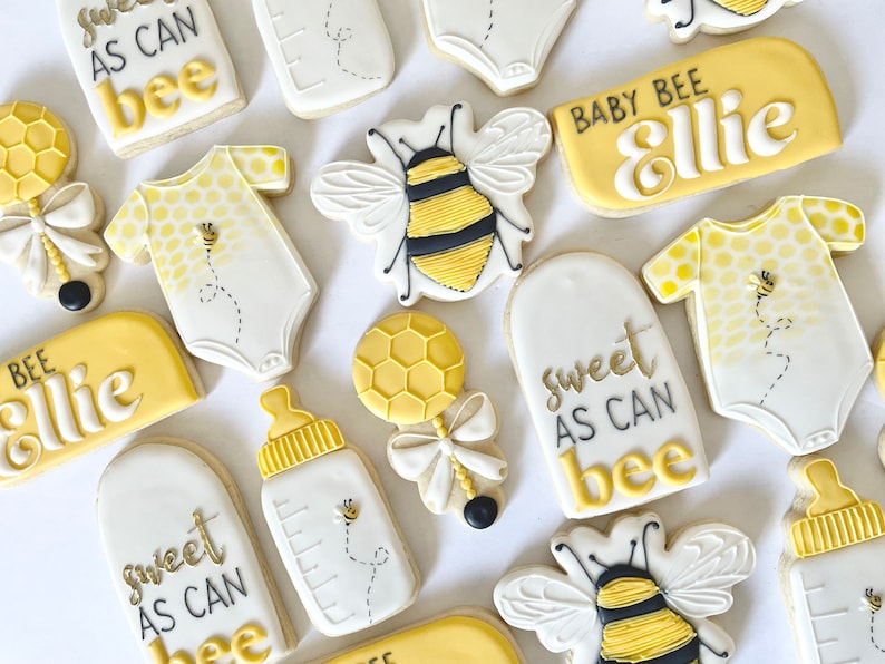 K&ouml;nnte beinhalten: Ein Set mit 10 gelben und wei&szlig;en, verzierten Zuckerkeksen mit Bienen- und Babymotiven. Die Kekse zeigen den Text "Sweet as can bee", "Baby Bee Ellie" und "Bee Ellie".