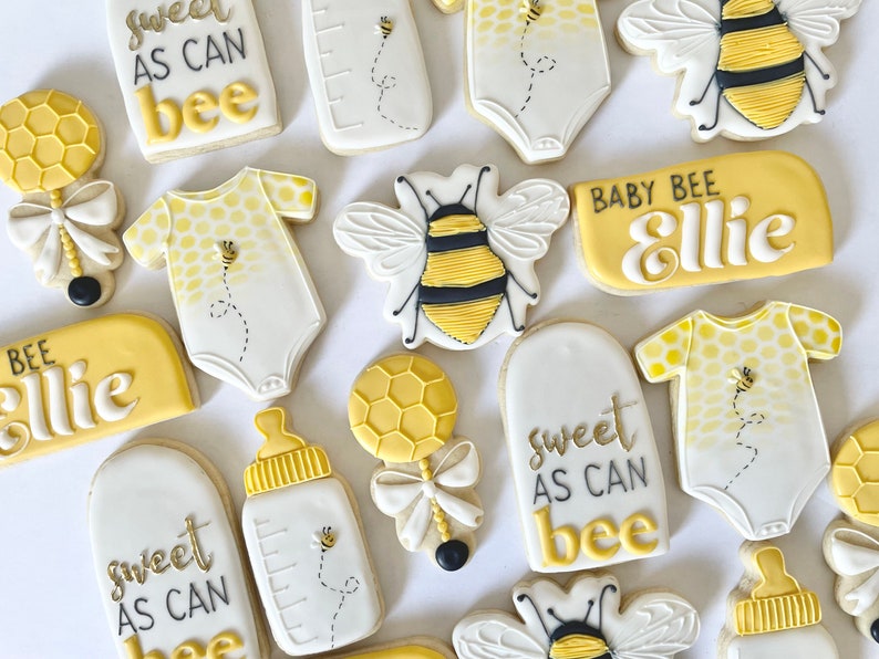 K&ouml;nnte beinhalten: Ein Set aus neun dekorierten Zuckerkeksen mit Bienen-Motiv. Die Kekse zeigen verschiedene Designs, darunter eine Babyflasche, eine Rassel, eine Biene und den Text "Sweet as can bee" und "Baby Bee Ellie".
