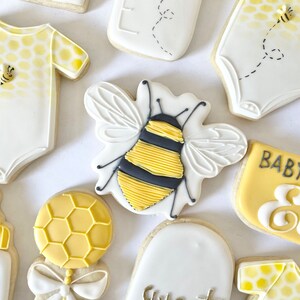 K&ouml;nnte beinhalten: Ein Set dekorierter Zuckerkekse mit einer Biene, einem Baby-Strampler, einer Flasche, einer Rassel und einem Schild mit der Aufschrift "Baby Bee Ellie".