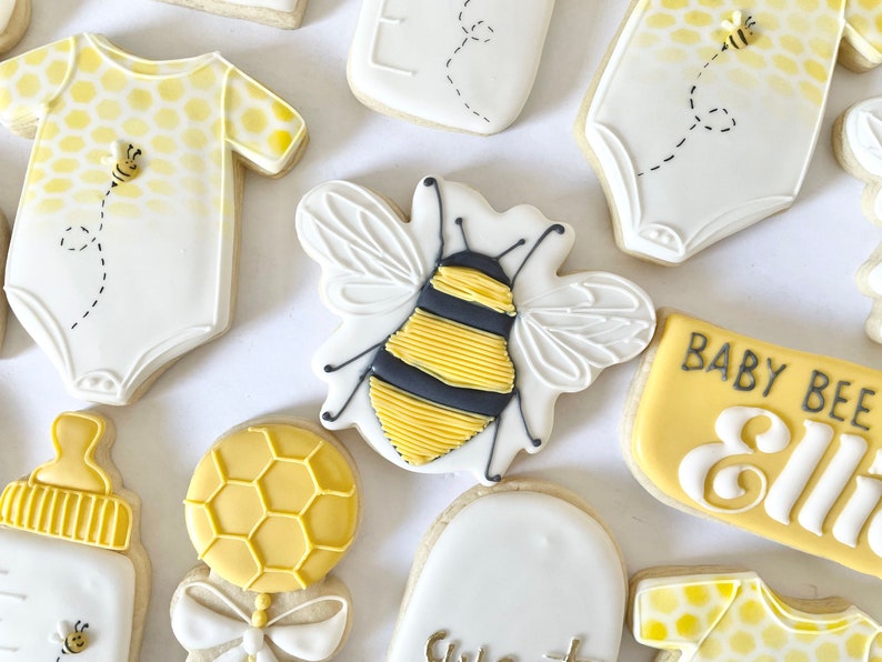 K&ouml;nnte beinhalten: Ein Set dekorierter Zuckerkekse mit einer Biene, einem Baby-Strampler, einer Flasche, einer Rassel und einem Schild mit der Aufschrift "Baby Bee Ellie".