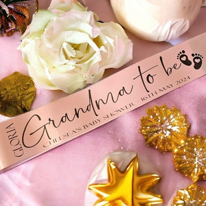 May include: A pale pink ribbon with the text "Grandma to be" in cursive, the date "16th May 2024", and baby footprints. The ribbon is on pink fabric, with a white rose, gold star decorations, and other decorative items. A baby shower theme.