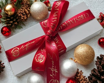 May include: A rectangular white gift box wrapped with a red satin ribbon. The ribbon features gold foil text that reads Merry Christmas and The Smiths, along with gold foil illustrations of Santa Claus faces tied in a decorative bow.