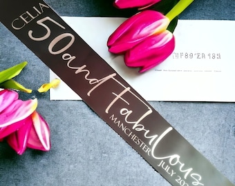 May include: A dark brown satin sash with the text "CELIA 50 and Fabulous MANCHESTER JULY 2023" in a light gold script. The sash is decorated with bright pink tulips and a white card.
