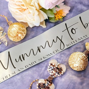 May include: A pale grey satin sash with the words "Mummy to be" in black script, along with "Chelsea's Baby Shower" and the date. The sash is surrounded by gold ornaments and flowers, ideal for a baby shower celebration.