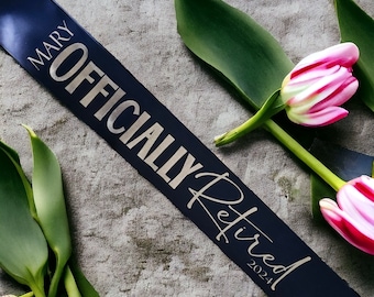 May include: A navy blue satin sash with silver text reading "MARY OFFICIALLY Retired 2024". The sash is laid diagonally on a textured grey surface, alongside pink and white tulips and green leaves.