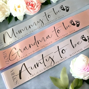 May include: Three satin baby shower ribbons in grey, pink, and white. Each ribbon has text that reads "Mummy to be", "Grandma to be", and "Aunty to be" with the date "16th May 2024". The ribbons are decorated with baby feet.