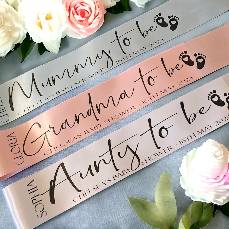 May include: Three satin baby shower ribbons in grey, pink, and white. Each ribbon has text that reads "Mummy to be", "Grandma to be", and "Aunty to be" with the date "16th May 2024". The ribbons are decorated with baby feet.