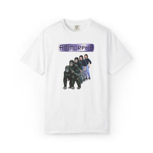 Puede incluir: Camiseta blanca con la palabra "ANIMORPHS" en un diseño rectangular morado y plateado. Debajo, un gráfico de un gorila transformándose en una persona. El diseño está centrado en la camiseta.
