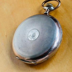 May include: Antique pocket watch with a silver-colored, textured face and a copper-colored ring and crown. The watch has a circular design with a detailed, engraved pattern. The watch is set against a light brown wooden surface.