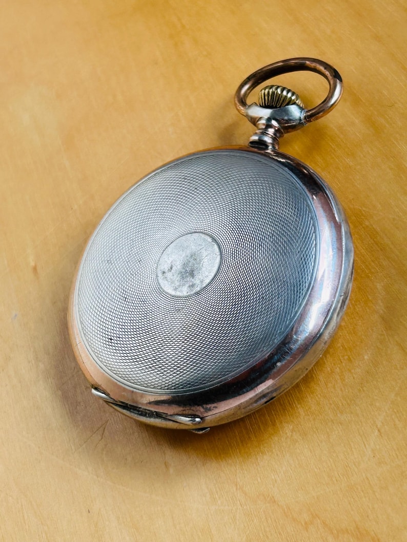 May include: Antique pocket watch with a silver-colored, textured face and a copper-colored ring and crown. The watch has a circular design with a detailed, engraved pattern. The watch is set against a light brown wooden surface.