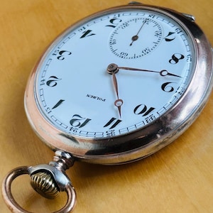 May include: Antique pocket watch with a white face, black numerals, and a subdial. The watch has copper-colored hands and a copper-colored case. The watch reads "POLHEM" on the face. The watch is an oval shape with a loop and a decorative crown.