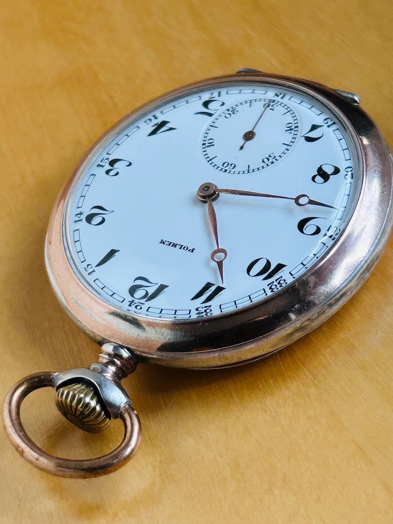 May include: Antique pocket watch with a white face, black numerals, and a subdial. The watch has copper-colored hands and a copper-colored case. The watch reads "POLHEM" on the face. The watch is an oval shape with a loop and a decorative crown.