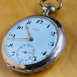 May include: A vintage pocket watch with a white face, black numerals, and a smaller seconds dial. The watch has a silver case with a copper-colored crown and a loop for a chain. The word "POLHEM" is printed on the face.