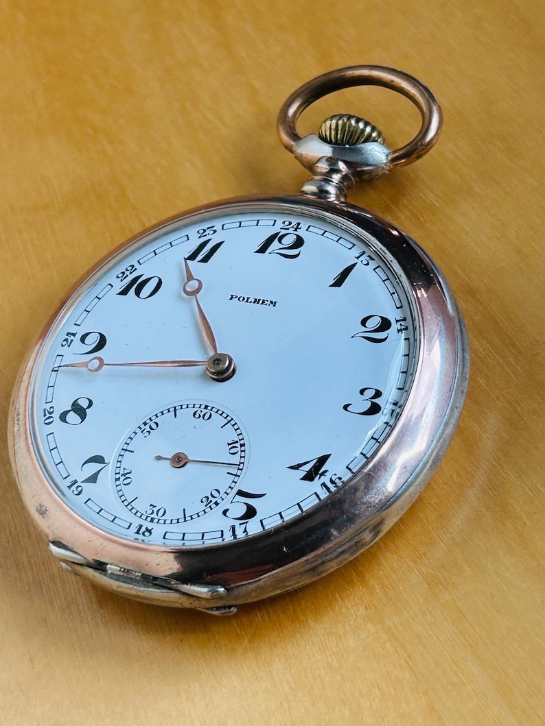 May include: A vintage pocket watch with a white face, black numerals, and a smaller seconds dial. The watch has a silver case with a copper-colored crown and a loop for a chain. The word "POLHEM" is printed on the face.