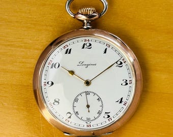 Peut inclure: Une montre de poche Longines vintage avec un cadran blanc, des aiguilles dorées et un sous-cadran. La montre a un boîtier doré et une tige de remontoir argentée. Le cadran affiche le logo Longines et des chiffres arabes.