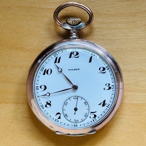 May include: A vintage pocket watch with a white face, black numerals, and a smaller seconds dial. The watch has a silver-colored case and a rose gold-colored crown and loop. The word "POLHEM" is printed on the watch face.