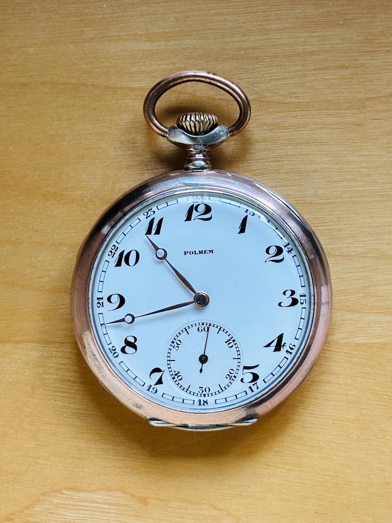 May include: A vintage pocket watch with a white face, black numerals, and a smaller seconds dial. The watch has a silver-colored case and a rose gold-colored crown and loop. The word "POLHEM" is printed on the watch face.