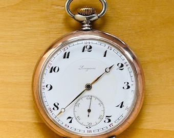 May include: A vintage Longines pocket watch. It features a white face with black numerals and a small seconds dial. The silver-tone case has a gold-tone crown and hands. The watch is displayed on a wooden surface.