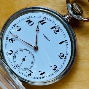 May include: A vintage pocket watch with a white face, black numerals, and copper-colored hands. The watch has a subdial for seconds and the brand name "POLHEM" is visible. The watch is open, revealing the face and the metal casing.