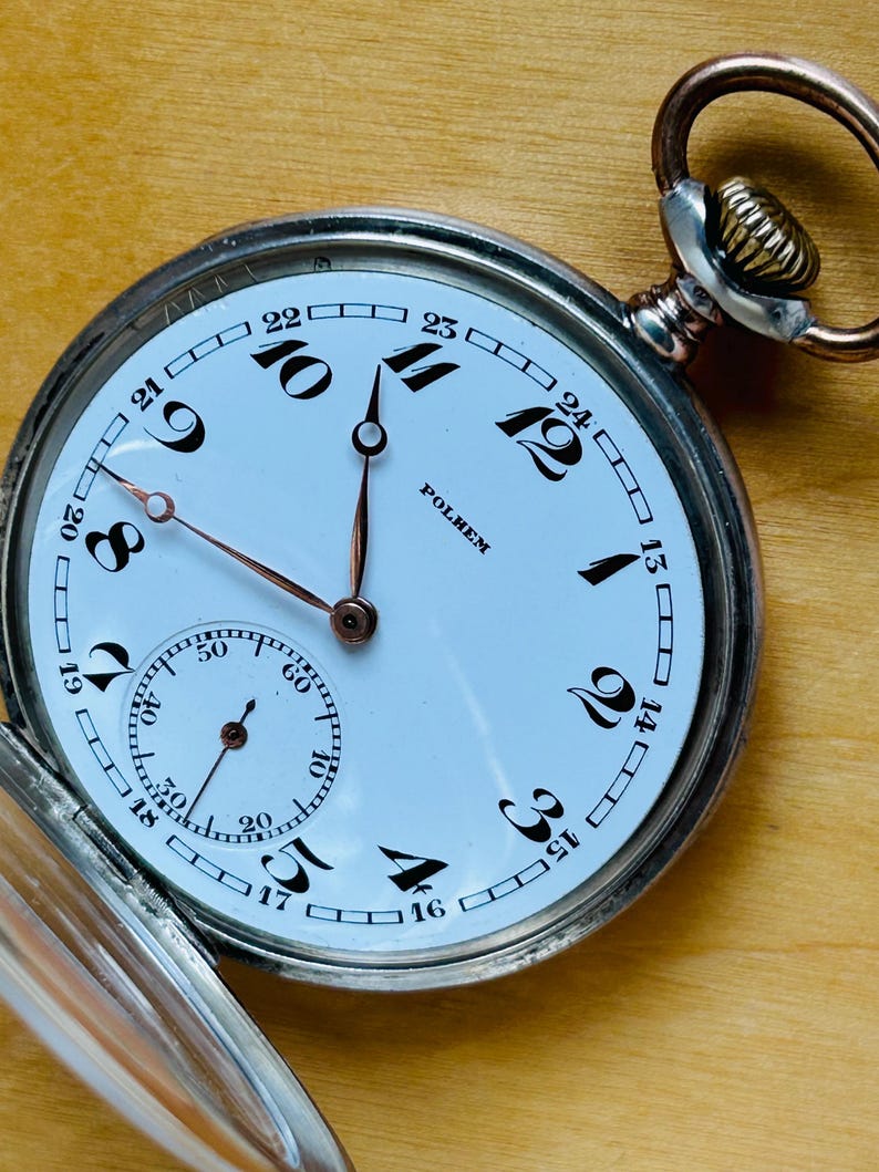 May include: A vintage pocket watch with a white face, black numerals, and copper-colored hands. The watch has a subdial for seconds and the brand name "POLHEM" is visible. The watch is open, revealing the face and the metal casing.