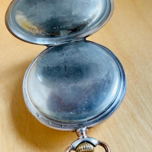 May include: An open, antique pocket watch with a silver-colored case and a gold-colored ring. The watch's interior is visible, showing a tarnished surface. The watch is resting on a light brown wooden surface.