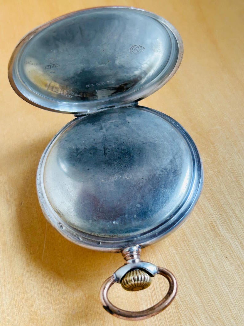 May include: An open, antique pocket watch with a silver-colored case and a gold-colored ring. The watch's interior is visible, showing a tarnished surface. The watch is resting on a light brown wooden surface.