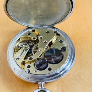 May include: Open silver pocket watch with the back lid ajar, revealing the intricate gold-colored gears and mechanisms. The watch has a silver case and a decorative loop for a chain. The serial number 070875 is visible on the inside of the back lid.