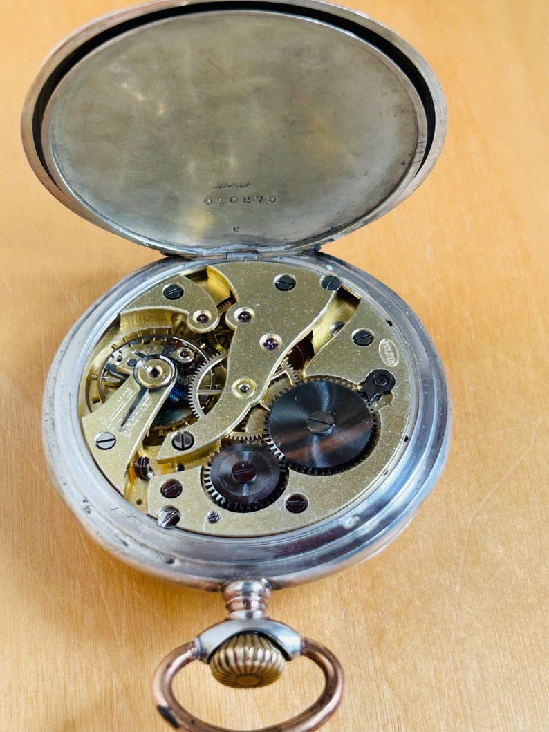 May include: Open silver pocket watch with the back lid ajar, revealing the intricate gold-colored gears and mechanisms. The watch has a silver case and a decorative loop for a chain. The serial number 070875 is visible on the inside of the back lid.