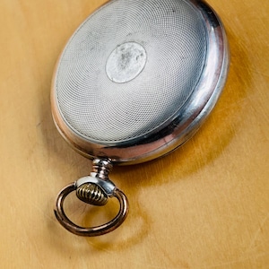 May include: Silver pocket watch with a detailed, textured face and a decorative ring. The watch has a circular design on the face and a gold-colored ring attached to the top. The watch is set against a light brown wooden surface.