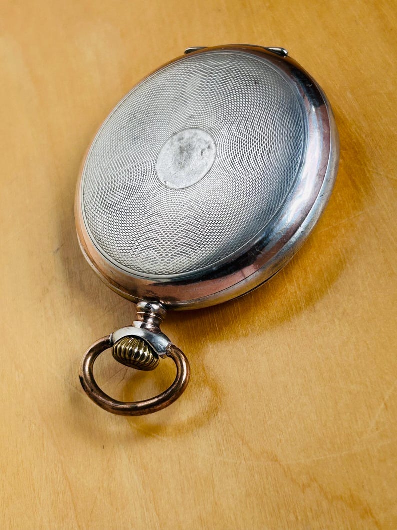 May include: Silver pocket watch with a detailed, textured face and a decorative ring. The watch has a circular design on the face and a gold-colored ring attached to the top. The watch is set against a light brown wooden surface.