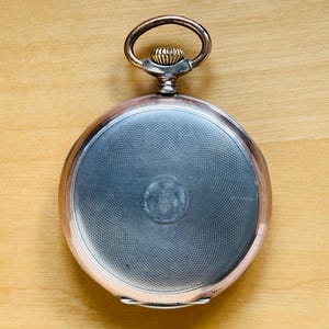May include: A vintage pocket watch with a silver-colored face and a copper-toned rim and loop. The watch face has a textured, circular pattern. The watch is set against a light brown wooden surface.