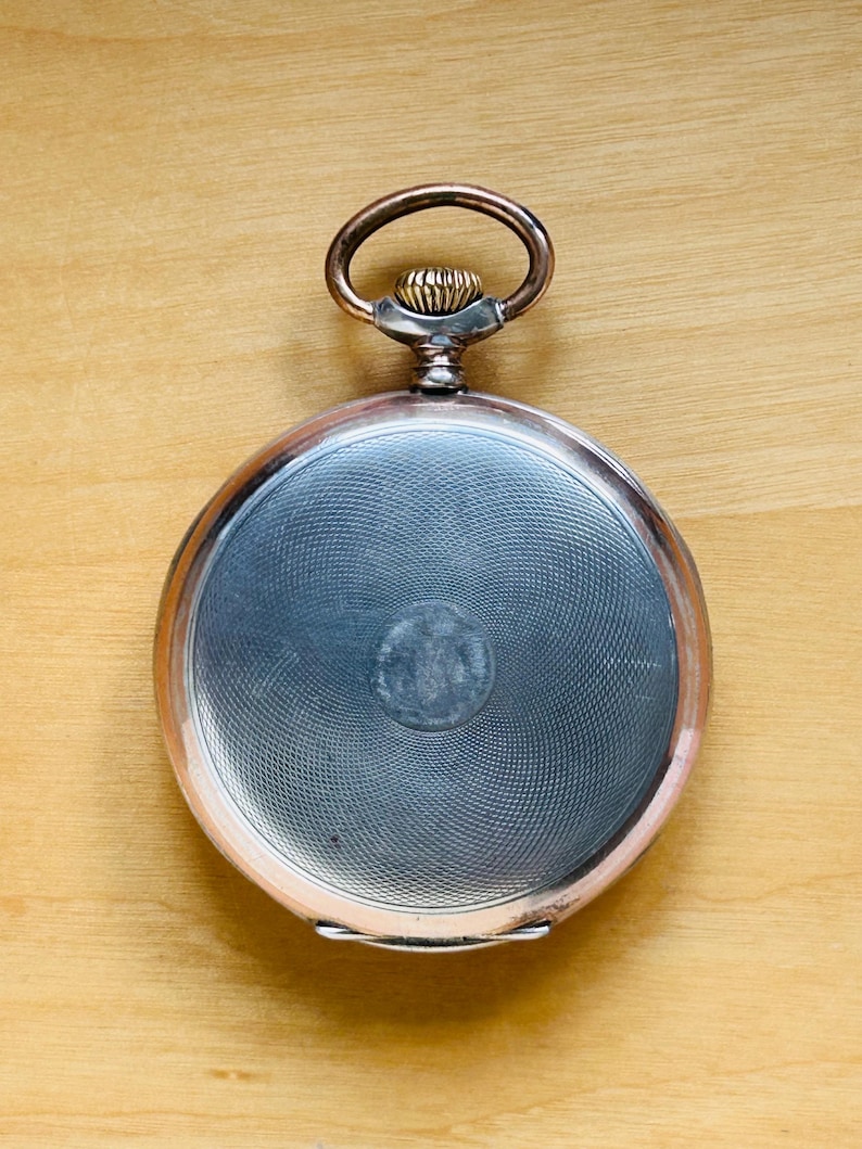 May include: A vintage pocket watch with a silver-colored face and a copper-toned rim and loop. The watch face has a textured, circular pattern. The watch is set against a light brown wooden surface.