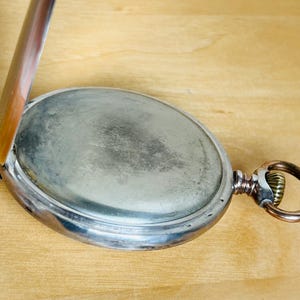 May include: An open, antique pocket watch with a silver-colored case and a tarnished, oval face. The watch's hinged cover is partially open, revealing the interior. A gold-toned ring and winding stem are visible on the side. The watch rests on a light brown wooden surface.