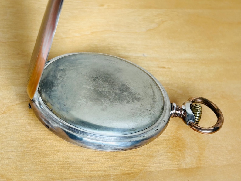 May include: An open, antique pocket watch with a silver-colored case and a tarnished, oval face. The watch's hinged cover is partially open, revealing the interior. A gold-toned ring and winding stem are visible on the side. The watch rests on a light brown wooden surface.