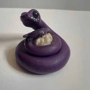 Peut inclure: Figurine de serpent violet enroulé autour d'un objet beige. Le serpent a des yeux noirs et des points blancs. Le petit serpent a des marques noires et blanches. La figurine est sur une surface blanche.
